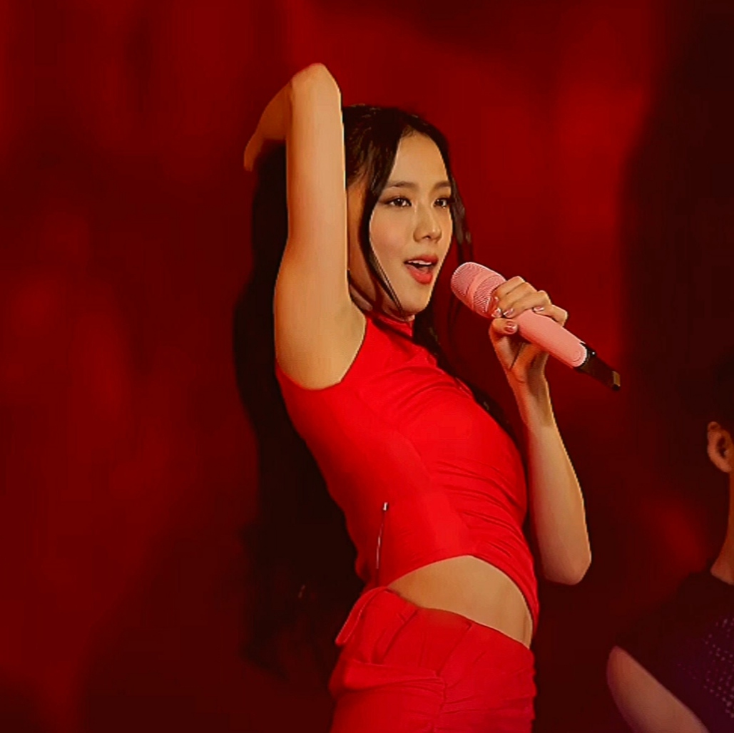 A woman in a red sleeveless crop top and matching pants sings into a pink microphone while raising her arm against a red backdrop.