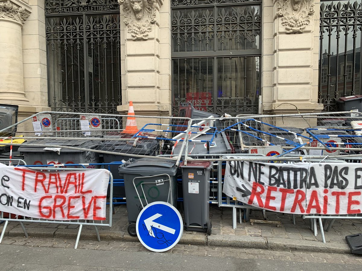 Autre angle de sciences po Lille bloqué. En plus des barrières poubelles et banderole on voit des panneaux de signalisation et des cônes de chantiers. Une autre banderole est attachée, on y lit " le travail on en grève"