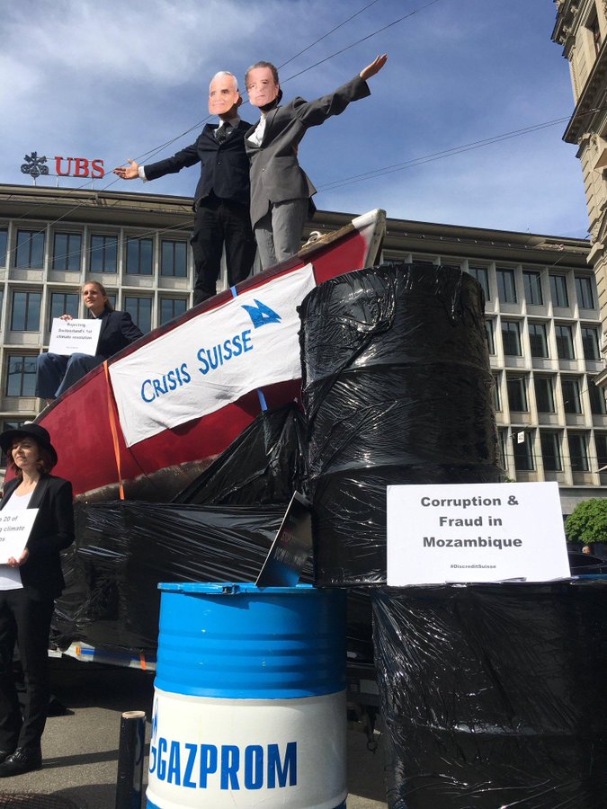 Zwei als Credit Suisse Manager verkleidete Personen auf einem Boot, beschriftet mit Crisis Suisse, das in Öl Fässer fährt. Im Hintergrund die UBS. 