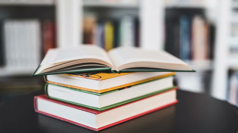 A pile of three books with a fourth lying open on top. Blurred in the background is a white bookcase with lots of books. Photo credit: Shutterstock
