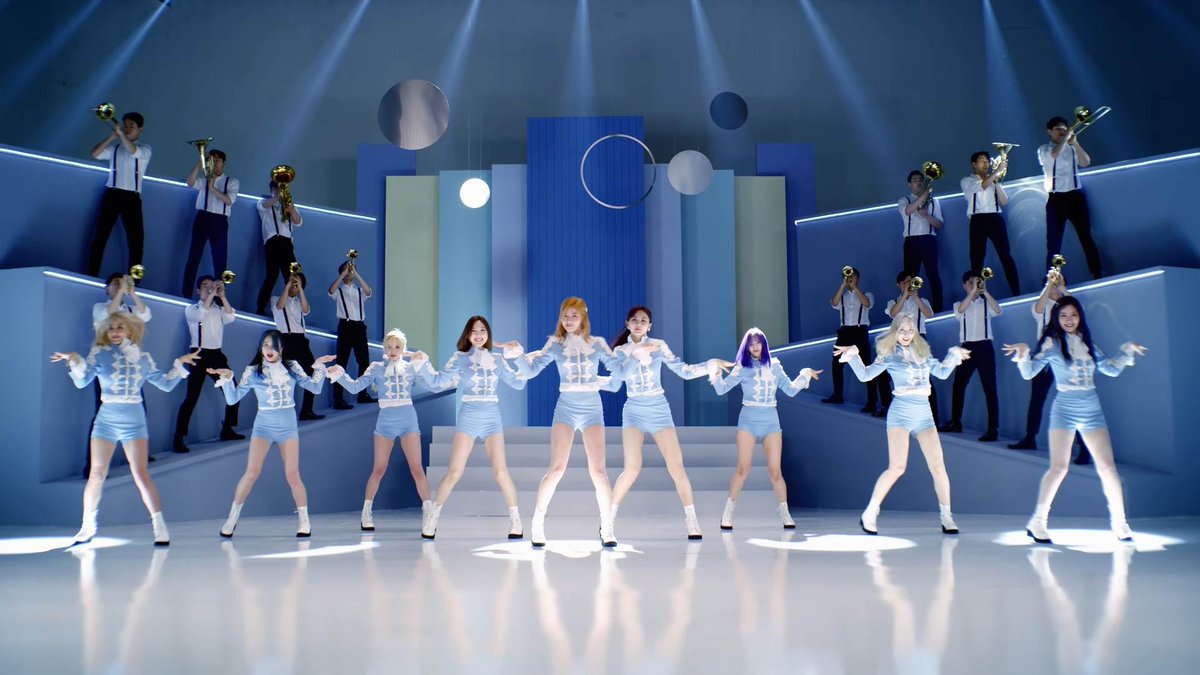A K-pop girl group performs a synchronized dance routine in light blue outfits with brass players behind them on a geometric blue and white stage with circular elements.