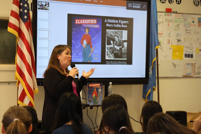 Traci Sorell, a light-skinned Cherokee woman with medium brown, shoulder-length hair, standing in a classroom in front of a projected image of the cover of Classified: The Secret Career of Mary Golda Ross, Cherokee Aerospace Engineer, which was illustrated by Natasha Donovan, and a photo of Mary with the heading "A Hidden Figure: Mary Golda Ross."