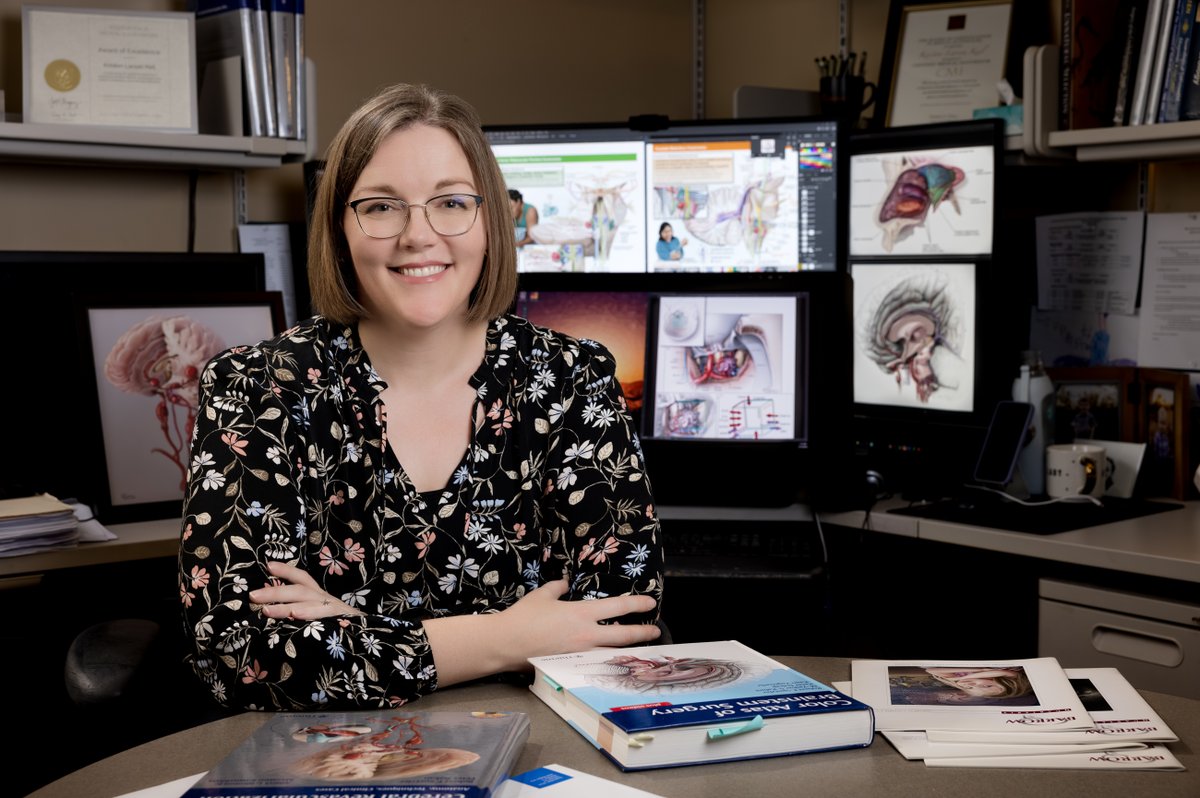 Kristen Larson Keil poses for a photo in front of her computer, where her medical illustrations are displayed on screens. She is leaning against a table, where more of her artwork is displayed in the form of textbooks and journals.