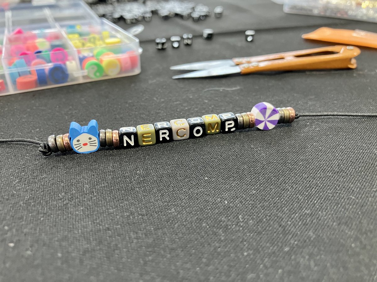 swkowk's tweet photo. Craft table at #NERCOMP23 (beaded bracelet spelling out NERCOMP with a blue cat bead at one end and a purple and white pinwheel bead at the other end) https://t.co/Fwp4mh4bi2