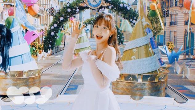 A young woman in a white dress smiling and waving in front of a festive, brightly decorated set with Christmas trees and balloons.