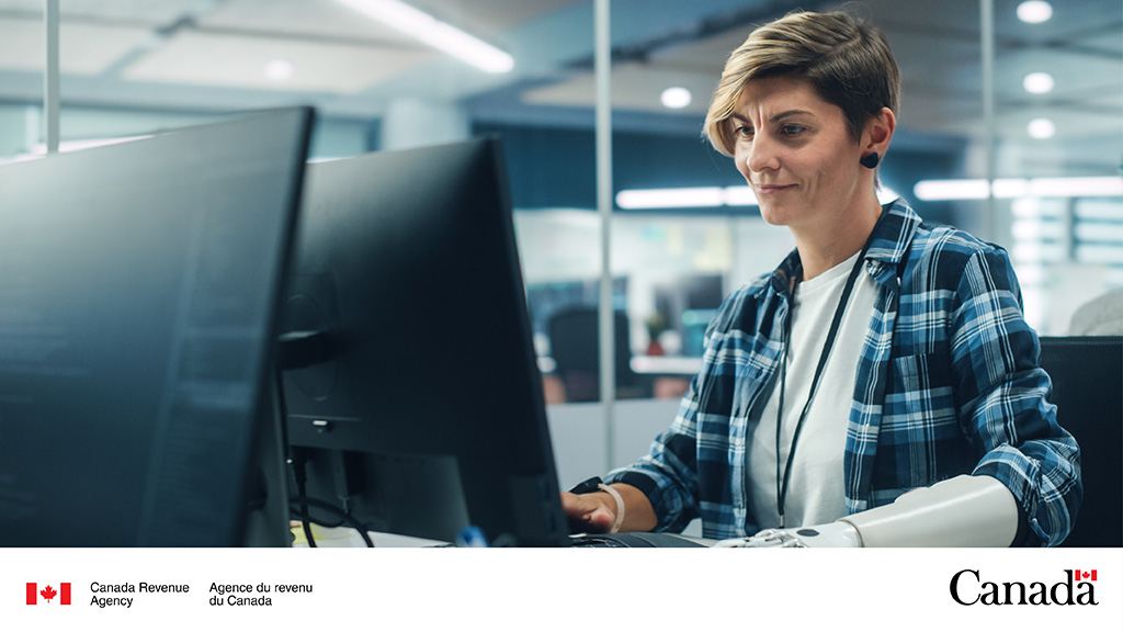 A person with short brown and blonde hair, wearing a blue plaid shirt, works at a desk in an office.