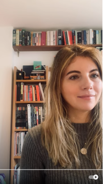 Photo of a smiling young white woman with long hair and wearing a grey jumper, looking at a computer screen just off to the right of the camera. There is a plain pale painted wall with bookshelves in front in the background.