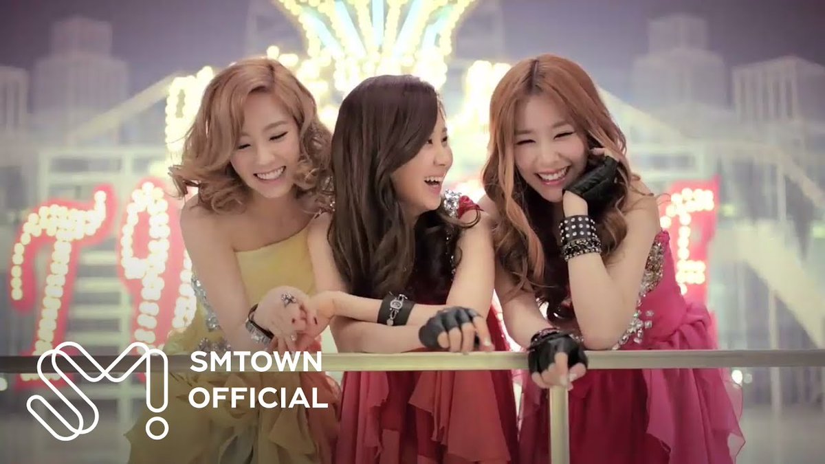 Three young women smiling and posing together in a colorful, glittery setting with bright lights in the background, featuring the 'SMTOWN OFFICIAL' logo, likely a K-pop music video scene.
