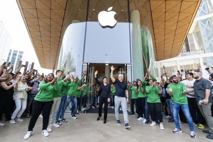 Tim and Deirdre stand and wave in front of the open doors at Apple BKC, surrounded by team members and customers.