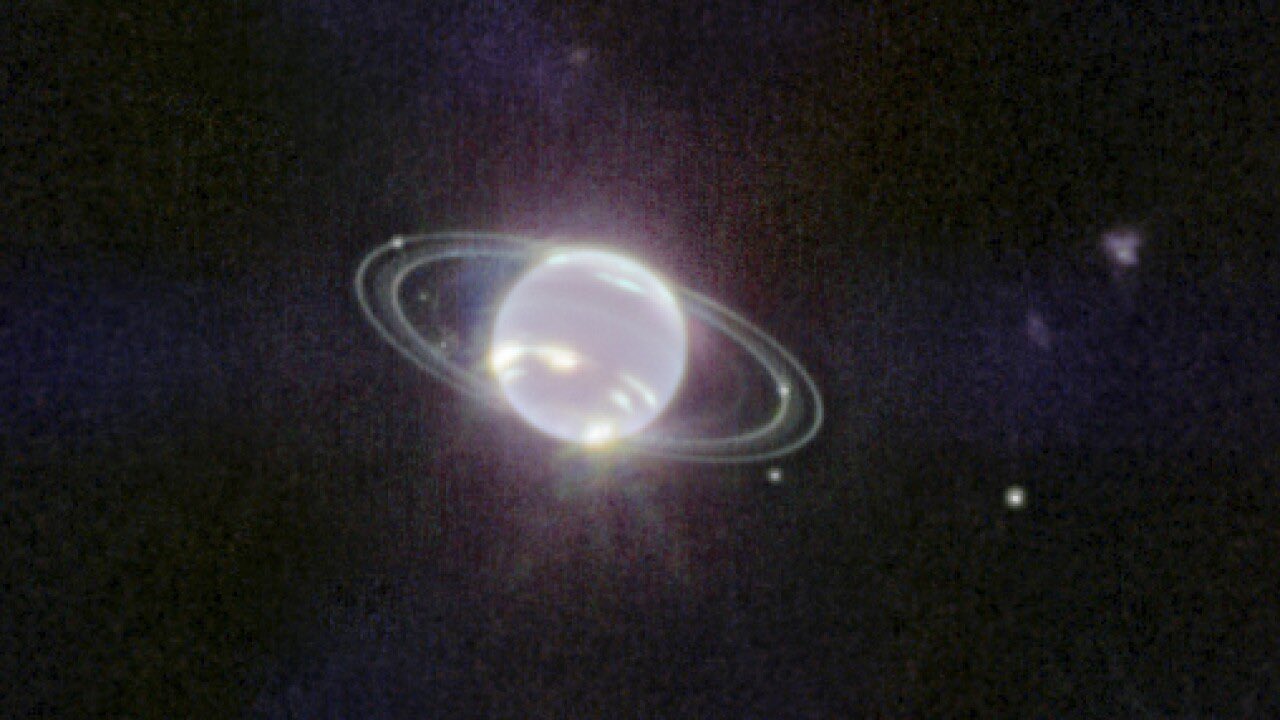 In this Webb image, Neptune resembles a pearl with rings that look like ethereal concentric ovals around it. There are 2 thinner, crisper rings and 2 broader, fainter rings. A few extremely bright patches on the lower half of Neptune represent methane ice clouds. Six tiny white dots, which are six of Neptune’s 14 moons, are scattered among the rings. The background of the image is black.