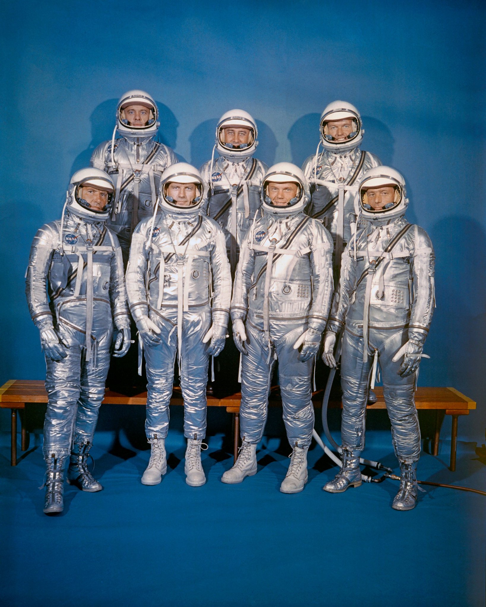 Seven astronauts in shiny Mercury spacesuits and helmets, arranged in two rows with three in the back and four in the front.