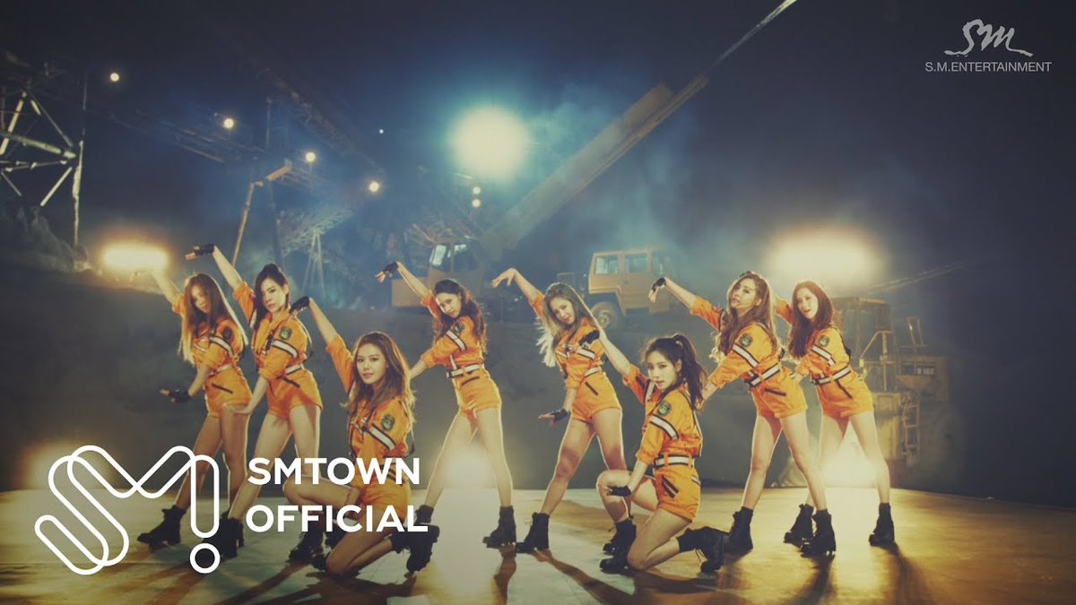 Nine female performers in orange outfits perform a choreographed dance on a stage with industrial equipment in the background, featuring SMTOWN OFFICIAL and S.M.ENTERTAINMENT logos.