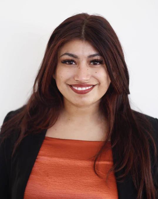 Headshot of Karina Hernandez against a white wall. She is smiling straight at the camera and is wearing an orange shirt with a black blazer. 