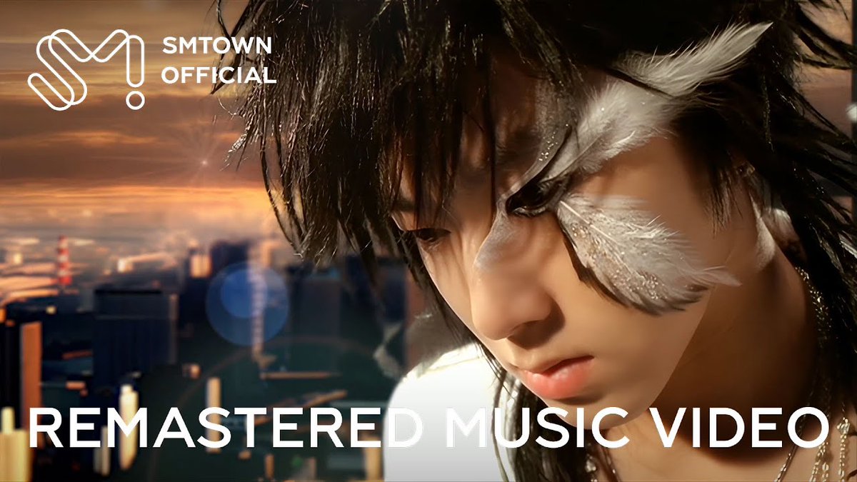 A thumbnail for an SMTOWN Official remastered music video, showing a close-up of a person with dark hair and white feather-like makeup around their eyes against a sunset cityscape.