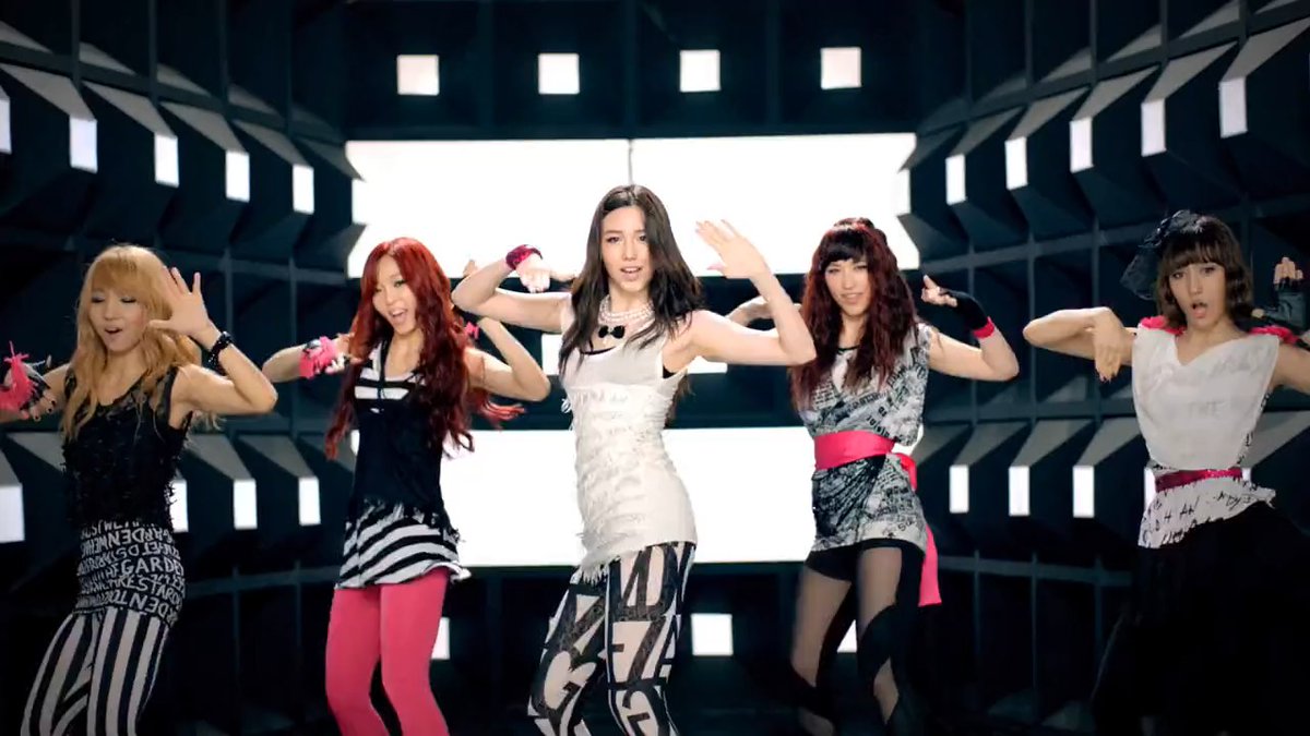 Five female K-pop performers dance in a coordinated routine on a stage with a black and white geometric backdrop, wearing contrasting black and white outfits with pink accents.