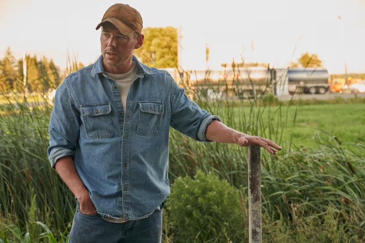 Brian J. Smith as Ken in Essex County. He stands in a lush farmers field, in practical denim farm-ware, leaning on a stake in the ground. 