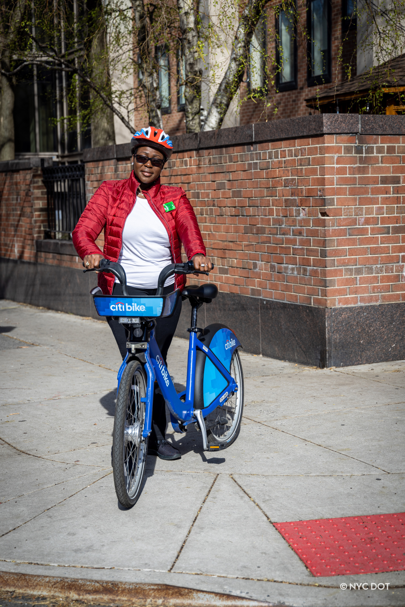 A person in a red jacket and a red white and blue helmet poses with a citi bike on a city sidewalk