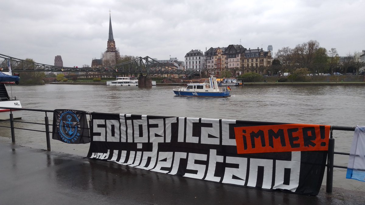 ein großes schwarzes Transpi hängt an einem Geländer am Main:<br>SOLIDARITÄT und WIDERSTAND immer!<br>links daneben die 'Scheisze Malaka Europa' Flagge von Iuventa Crew<br>im Hintergrund auf dem Main ein Polizeiboot vor dem Eisernen Steg