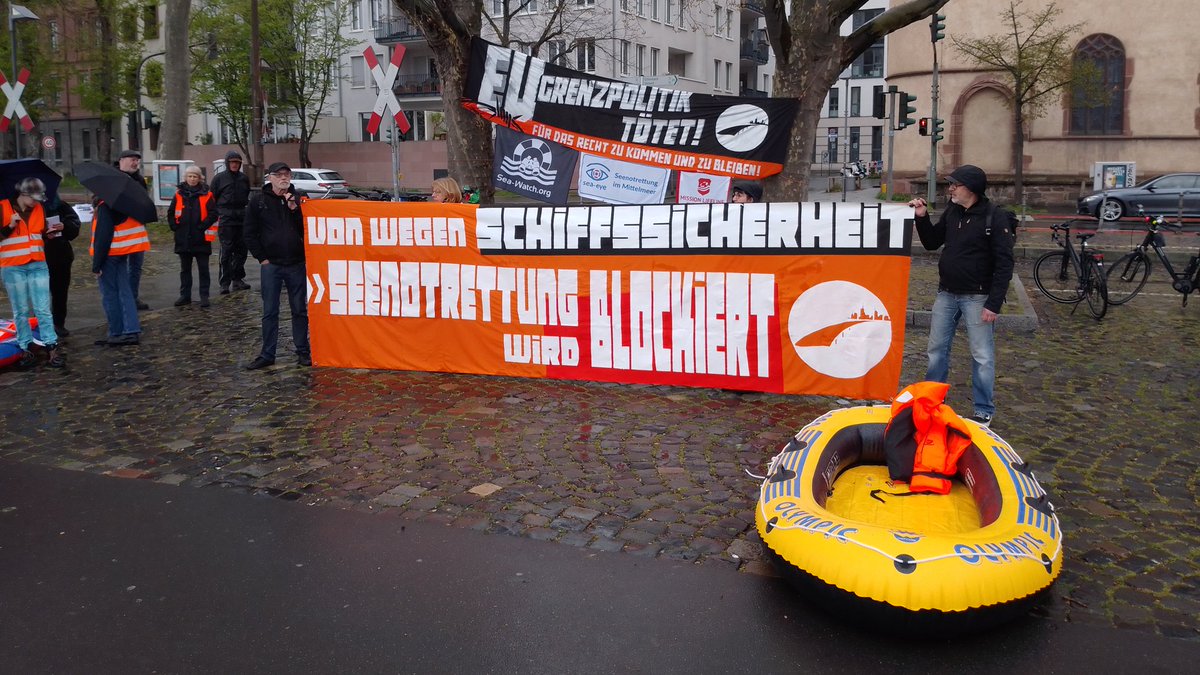 vorne ein Schlauchboot mit Rettungsweste;<br>dahinter stehen Menschen mit einem großen orangen Transparent:<br>von wegen SCHIFFSSICHERHEIT<br>SEENOTRETTUNG wird BLOCKIERT<br>daneben stehen Menschen mit orangen Sicherheitswesten<br>dahinter hängt zwischen Bäumen ein großes schwarzes Transpi:<br>EU GRENZPOLITIK TÖTET<br>darunter Flaggen von Sea Watch, Sea Eye & Mission Lifeleine