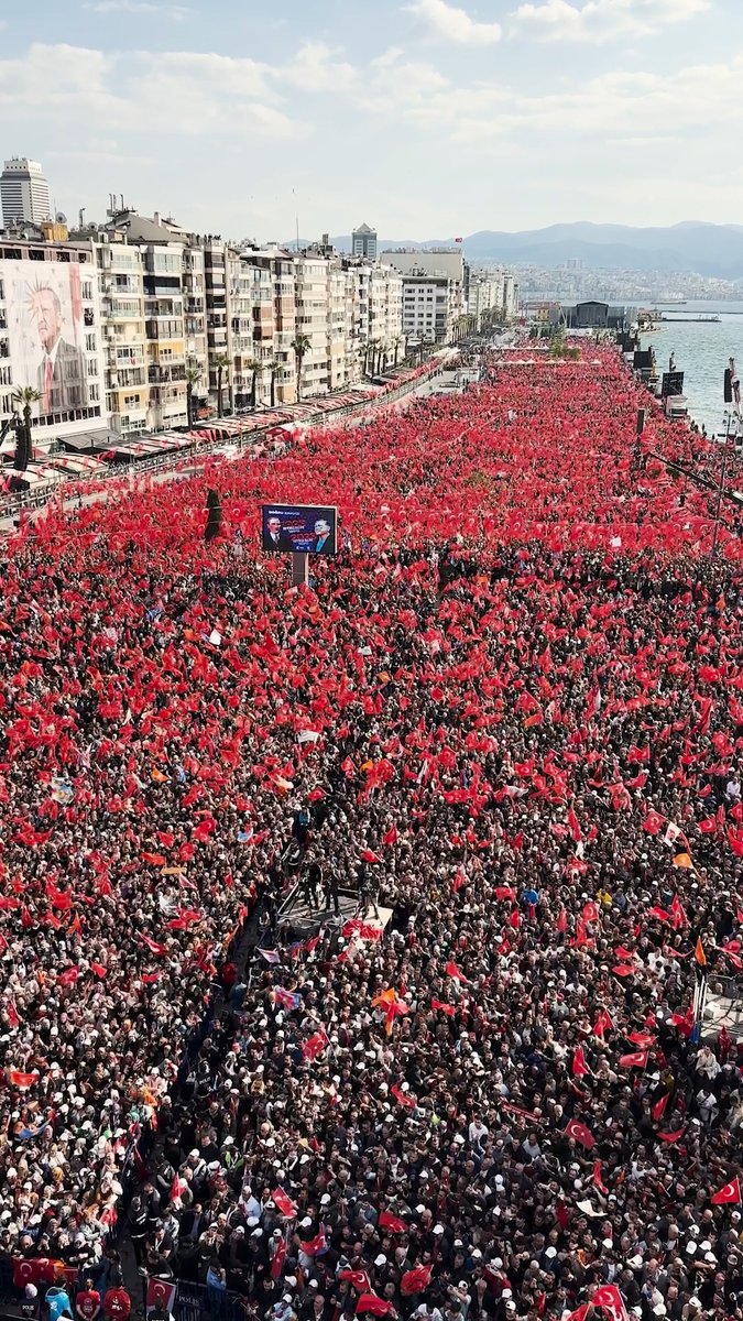 An aerial view captures a massive crowd of people filling a wide street along a waterfront, waving numerous Turkish and red flags.