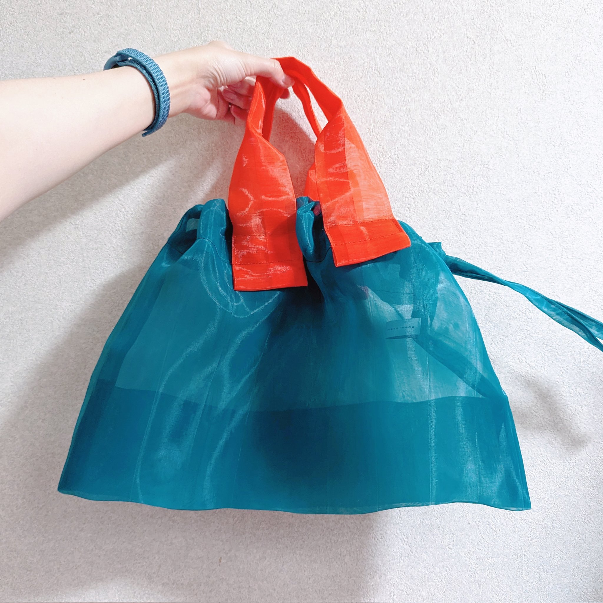 小松マテーレのすけるバッグマルチカラーを持ってる写真。カバン本体はピーコックカラーで、持ち手は鮮やかなオレンジ。