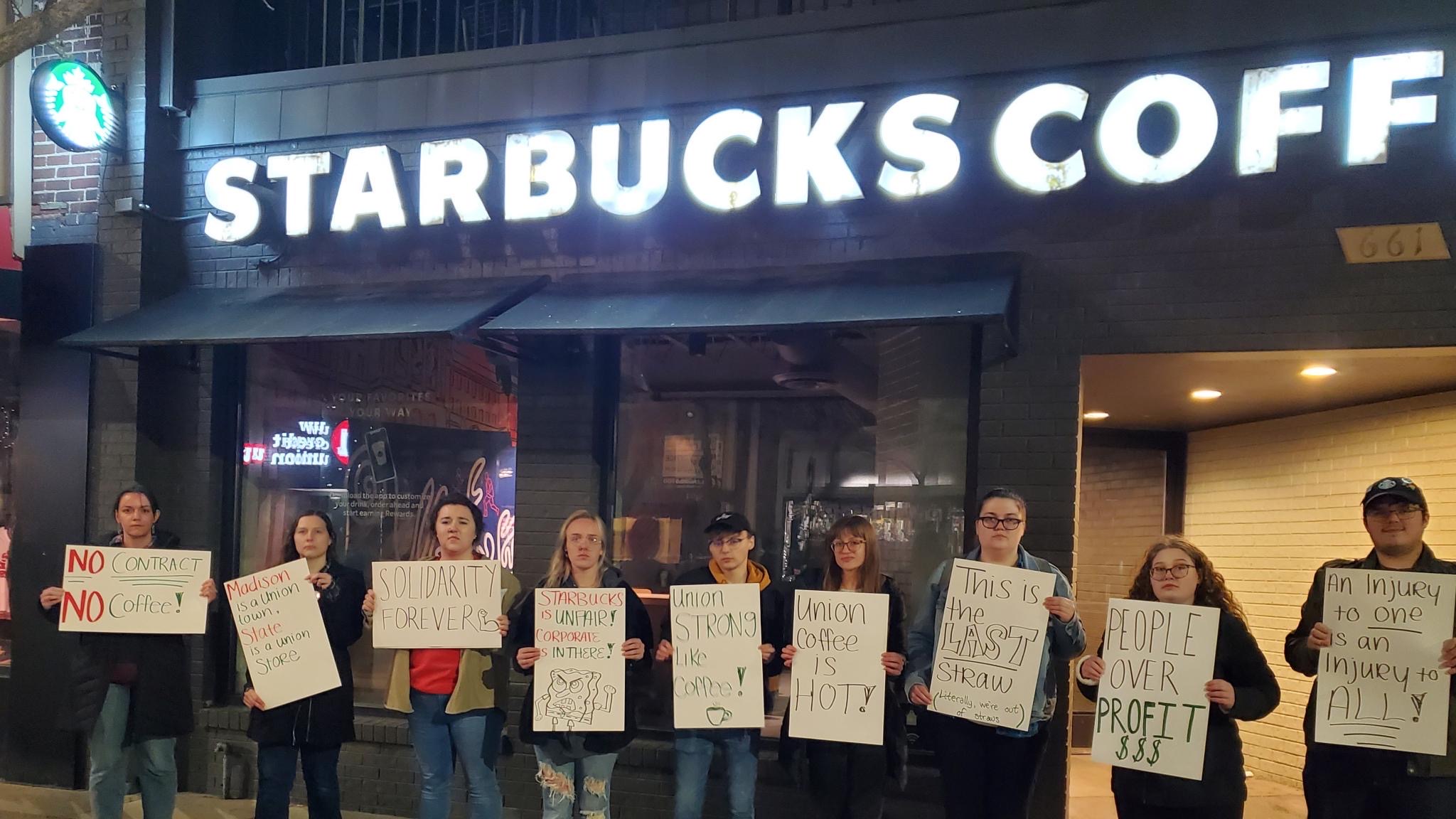 Nine Starbucks partners hold signs in front of their store in Madison, WI. Text on signs include "An injury to one is an injury to all!," "Union coffee is hot!", "People over profits," and "Starbucks is unfair! Corporate is in there!" next to a drawing of SpongeBob.