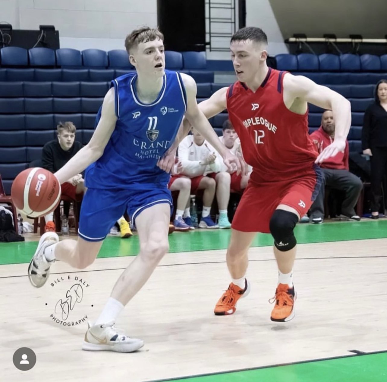 Basketball player in blue running past player in red whilst bouncing a basketball.