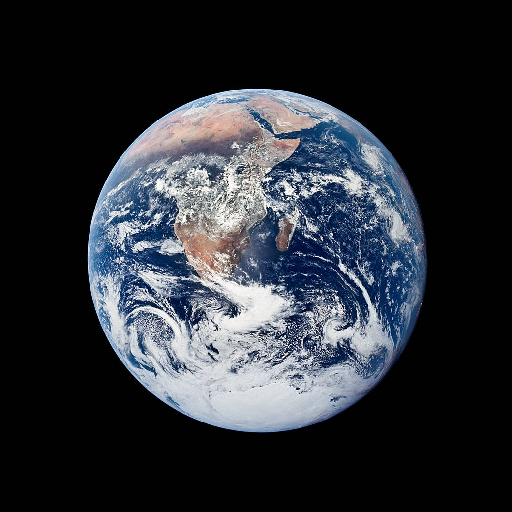 An image of the Earth, a round ball, set against the blackness of outer space. Through swirling clouds, the continent of Africa and the island of Madagascar can be seen; Antarctica is just visible at the very bottom of the Earth, and the Arabian peninsula at the top. The Atlantic and Indian Oceans are a deep, rich blue, giving this photo its common name: the Blue Marble.