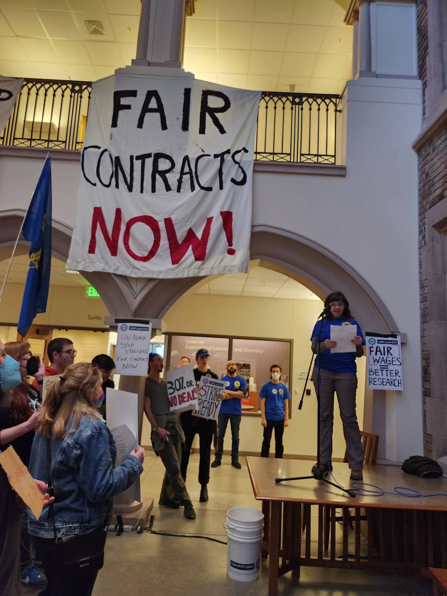 A UW researcher speaking in front of a crowd, with a sign that says "FAIR CONTRACTS NOW!" in the background 