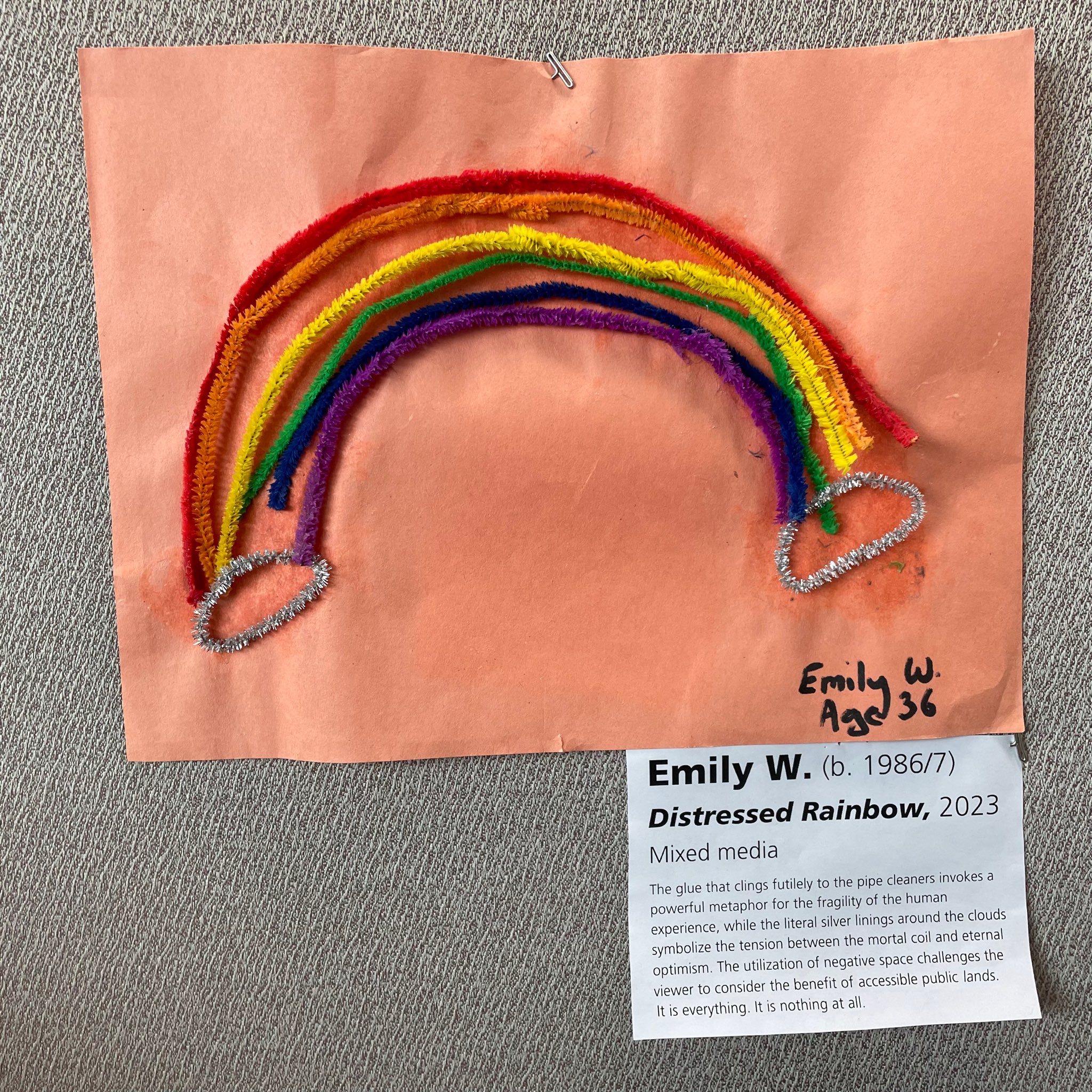 A rainbow composed pipe cleaners is glued to a sheet of orange construction paper. It is signed “Emily W. Age 36.” A mock-art gallery label is pinned underneath. It reads:
Emily W. Born 1986 or 1987.
Distressed rainbow, 2023
Mixed media
The glue that clings futilely to the pipe cleaners invokes a powerful metaphor for the fragility of the human experience, while the literal silver linings around the clouds symbolize tension between the mortal coil and eternal optimism. The utilization of negative space challenges the viewer to consider the benefit of accessible public lands. It is everything. It is nothing at all