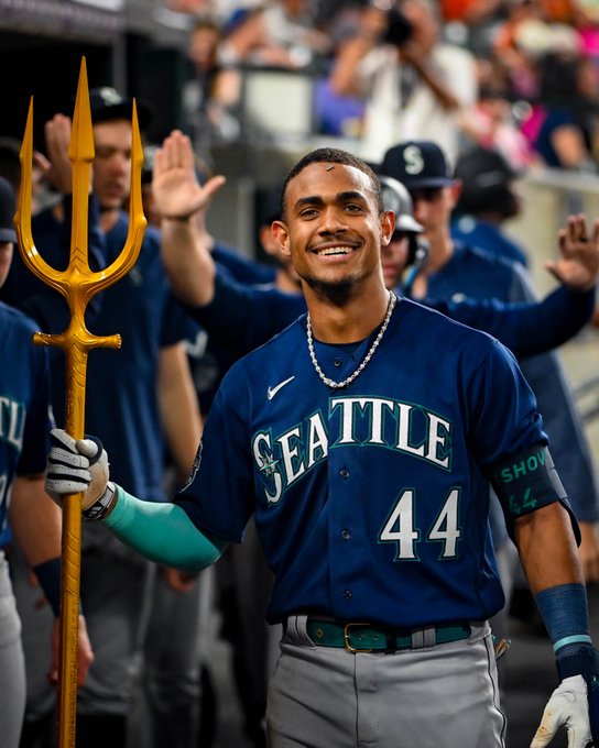 Julio Rodríguez, holding the home run trident, smiles at the cameras near the end of the dugout.
