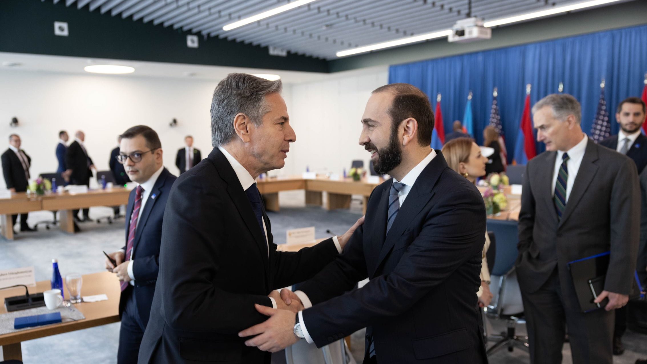 Secretary of State Antony Blinken shakes hands with Armenian Foreign Minister Ararat Mirzoyan in a large meeting room. Behind them are members of their delegations.