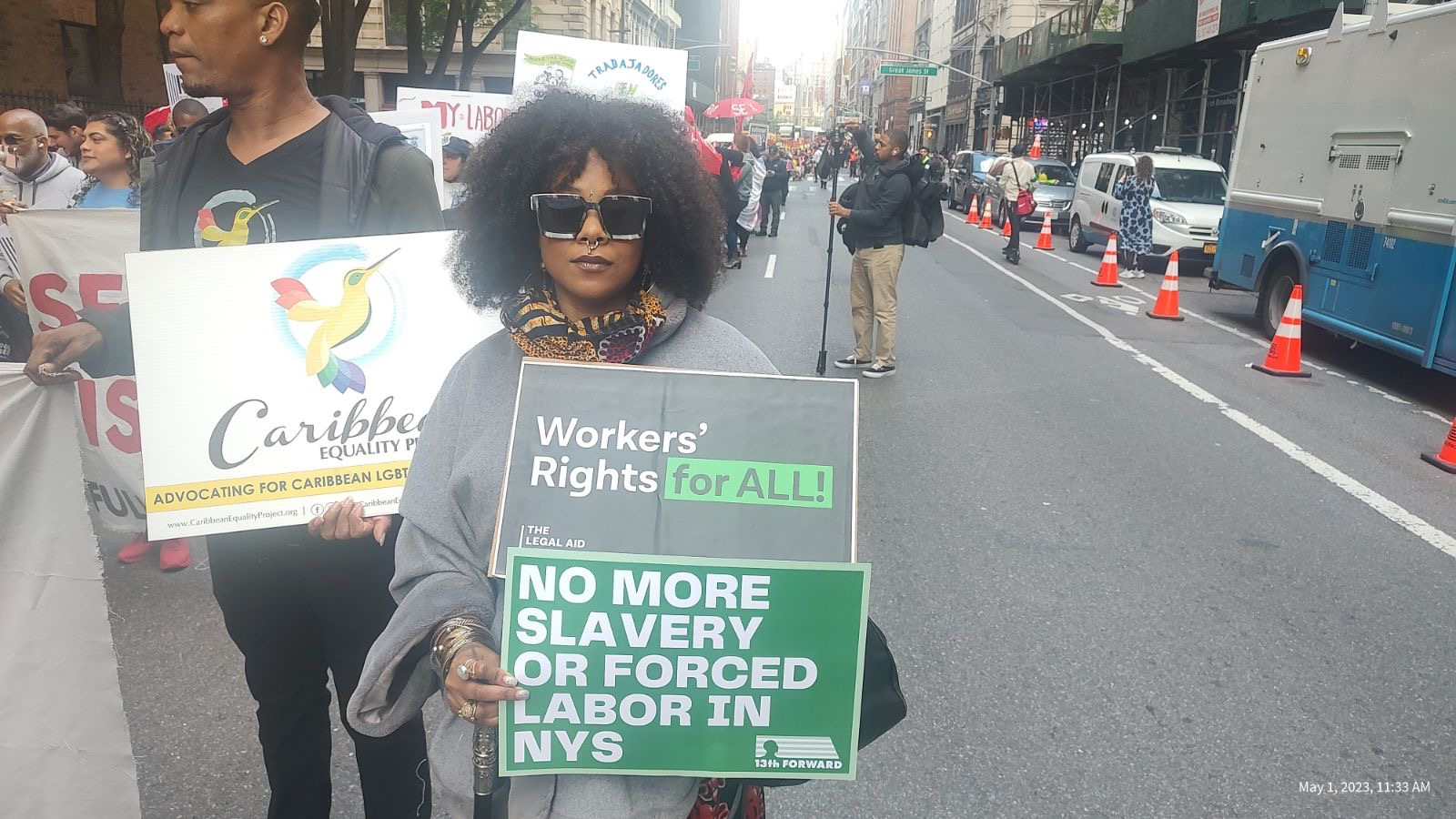 A person with curly hair, a nose ring and sunglasses in a crowd holding two signs. One says: “Workers’ Rights for ALL” and the other says “NO MORE SLAVERY OR FORCED LABOR IN NYS.” The first sign is from the Legal Aid Society and has their logo on it, and the second sign has the 13th Forward logo on it. The first sign is black with white text and the “for ALL” highlighted in green. The second sign is green with white text in all caps.