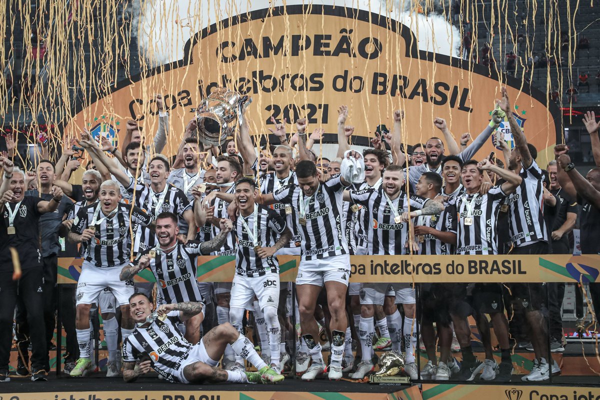 CopaDoBrasilCBF's tweet photo. 2021, o ano em que o @Atletico acabou com a emoção no futebol brasileiro. Ou melhor, monopolizou-a para si e para a massa: não tinha graça (ou chance) para mais ninguém. No Brasileirão, chegou ao topo na 15ª rodada e de lá nunca mais esteve nem perto de sair. Aos rivais, restava apenas brigar com a matemática. Bradar um “ainda dá” e, num esforço pueril, pensar numa combinação mirabolante de resultados e jogos a menos que pudesse, talvez, reduzir a vantagem a um confronto direto. Nunca aconteceu. 24 rodadas na liderança com uma média de 8 pontos de vantagem. Na que importa, a última, foram 13. O Galo sobrou demais o ano inteiro, voou solo em território nacional e, na minha disputa, não foi diferente. Exceto, talvez, pelas oitavas de final. O Atlético se viu precisando de um gol na Bahia para sacramentar sua classificação da mesma maneira que precisaria dele, meses depois, para concretizar sua tão esperada festa do título. O mesmo estado, o mesmo Esquadrão de Aço pelo caminho. Aos 55 do primeiro tempo, Juninho Capixaba estufava a rede, o Bahia devolvia os 2x0 e zerava a vantagem construída no Mineirão. Mas a agonia durou pouco. Vargas, aos 18 da segunda etapa, recolocou o Galo na frente. Dali em diante, nos placares agregados, 3x1 sobre o Fluminense nas quartas, 6x1 sobre o Fortaleza nas semis e outros 6x1 sobre o Athletico na final. Ganhou por 2x1 como visitante nos três confrontos… a caminhada foi quase um passeio no parque. Tal qual parecia, rodada a rodada, a trajetória no Brasileirão. Os jogos contra o Bahia foram os únicos em que os rivais puderam acreditar, por alguns minutos, no que eles mesmos diziam. O gol de Keno lá, o gol de Vargas aqui e definitivamente não dava mais nada. Cinquenta anos de uma piada repetida à exaustão jogados no lixo em apenas um. O Galo não tem um bi? É verdade, tem dois: bicampeão brasileiro e da #CopaBetanodoBrasil em 2021. Não apenas BI, não apenas CAMpeão brasileiro como SUPERcampeão brasileiro, de fato e de direito, naquela temporada. Novamente, a sensação generalizada é de que nem precisava de mais um jogo para que os atleticanos pudessem ostentar esse título… mas teve. E, adivinha? Ai, credo, o Galo ganhou mais uma vez! 🐓