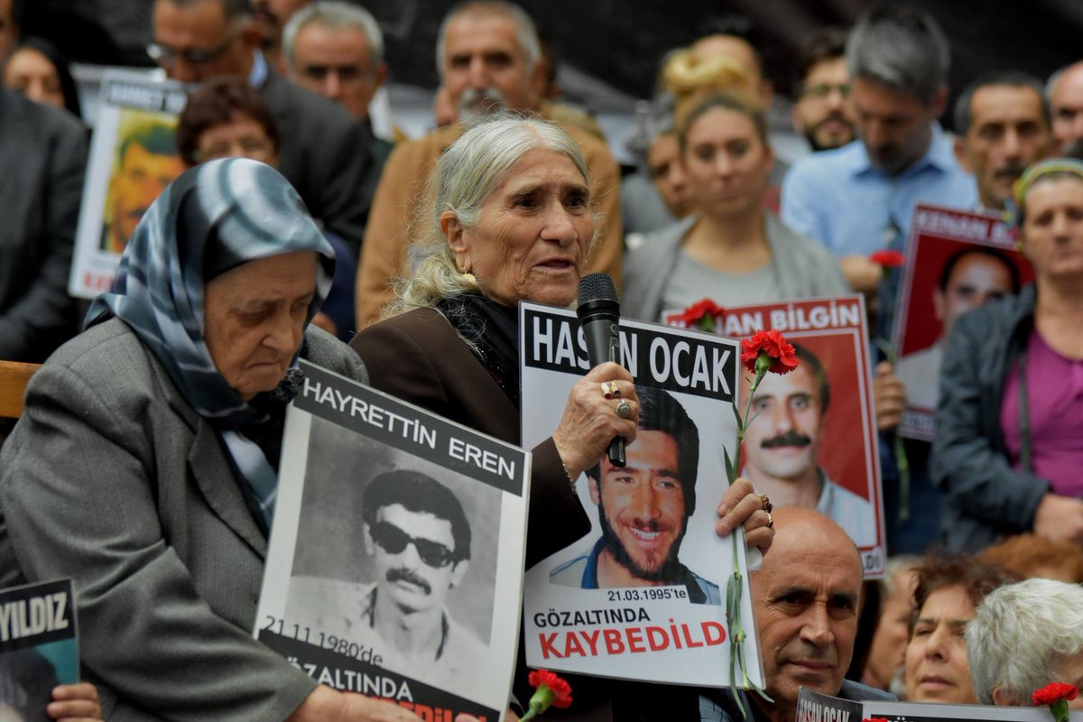 A crowd of women and men are holding signs with photos of victims of enforced disappearances. An old woman is speaking through a microphone.