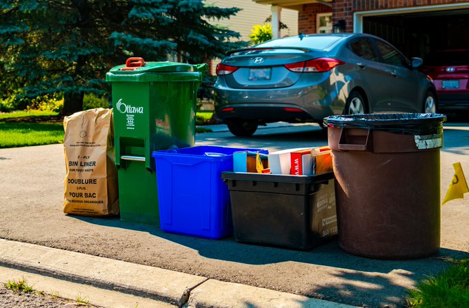 From left to right, a paper bin liner, a green bin, a blue bin, a black bin and a regular waste bin are lined up in the front of a driveway ready for collection.