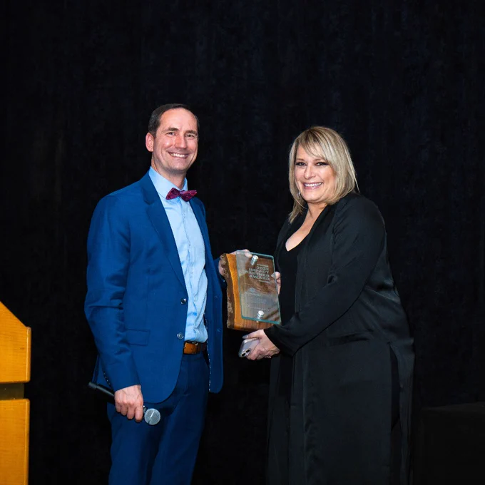 2 people standing in front of a black curtain at Mission Possible's 2022 Gala. They are dressed formally and are smiling at the camera while holding a glass plaque between them.