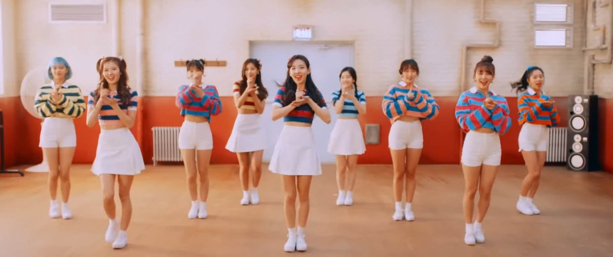 Nine female performers in colorful striped crop tops and white skirts dance in a bright rehearsal studio with orange lower walls.
