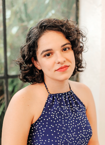 Foto da autora. Mulher jovem e branca. Possui cabelo preto e cacheado, na altura do queixo. Olha de lado para a câmera, com um leve sorriso. Veste uma regata azul.