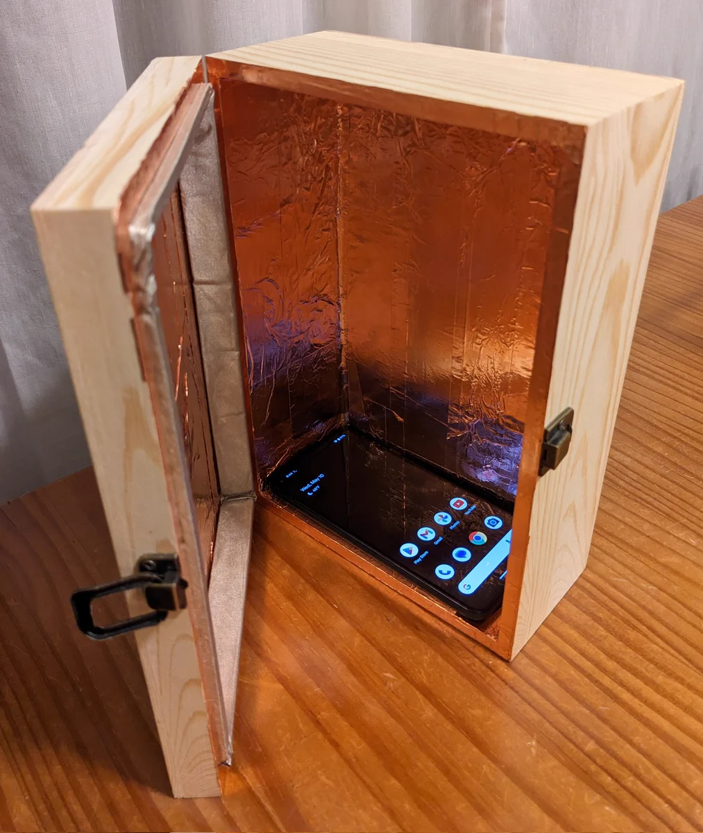 Photo of a mobile phone in a small pine box lined in copper foil tape and conductive fabric+foam.