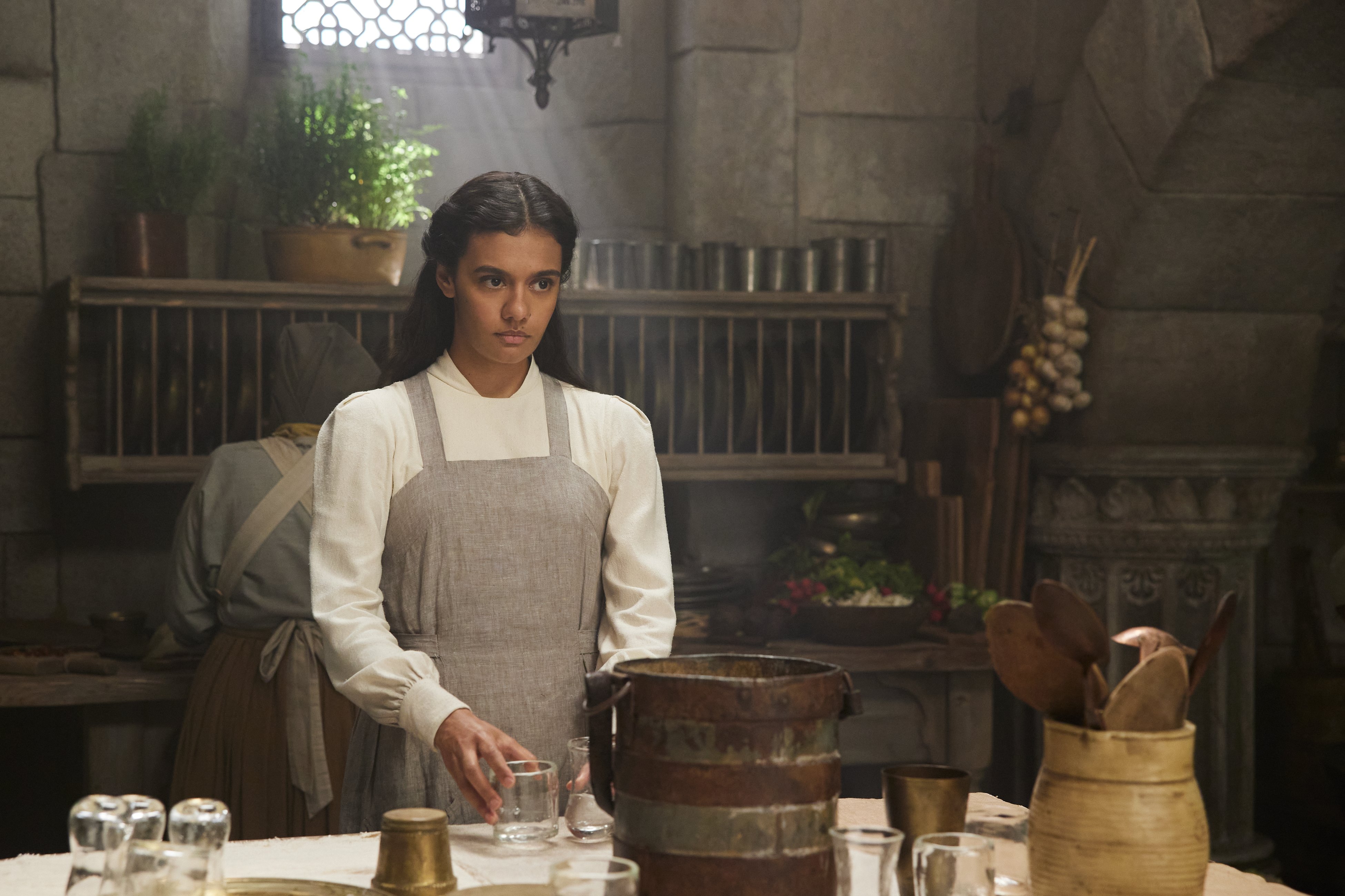 Medium shot of Egwene in a kitchen, holding a glass and small pitcher of water.