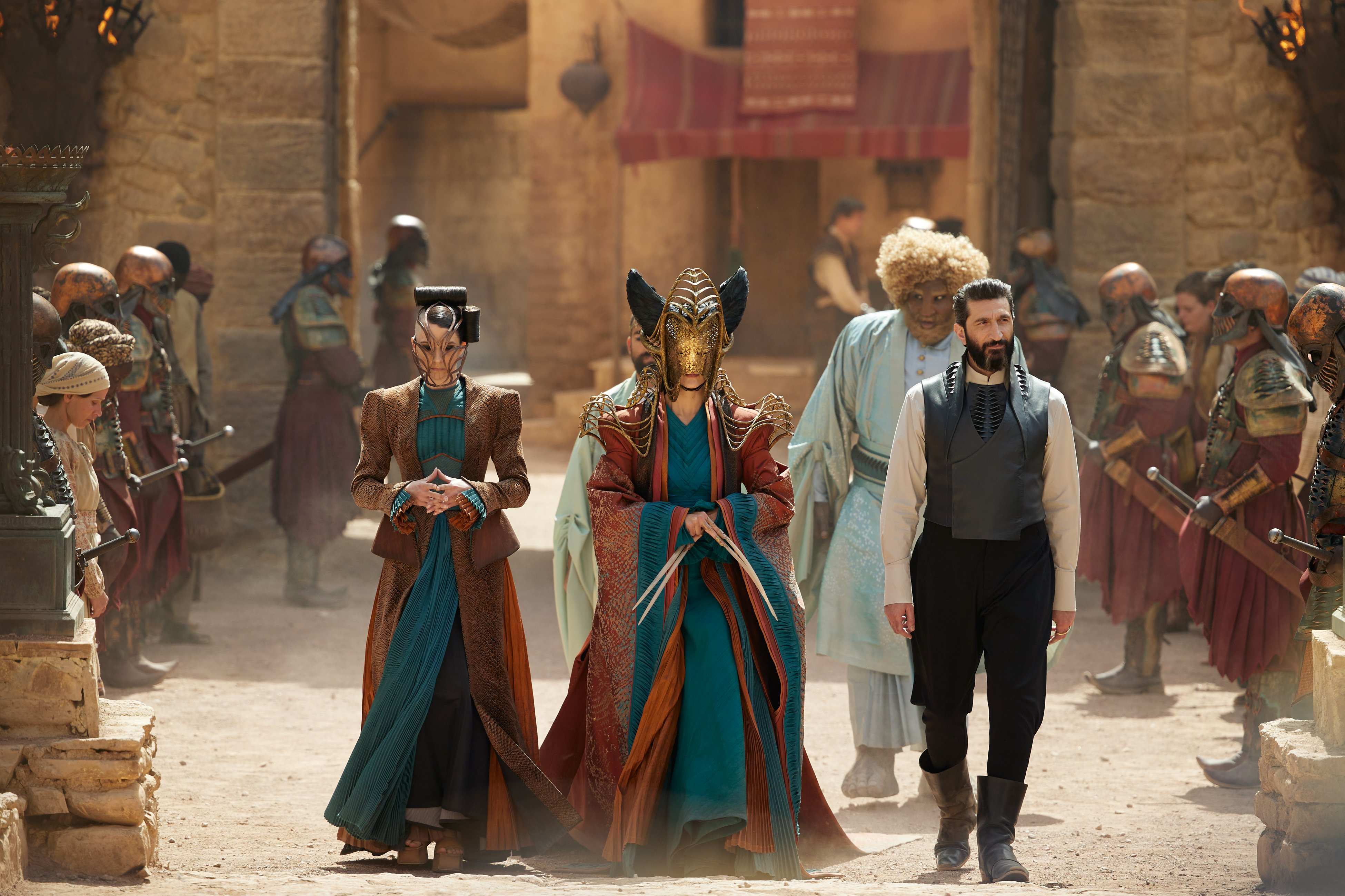 Full shot of five figures walking in formation, watched by a crowd. The central figure wears an ornate metal headpiece and has very long nails.