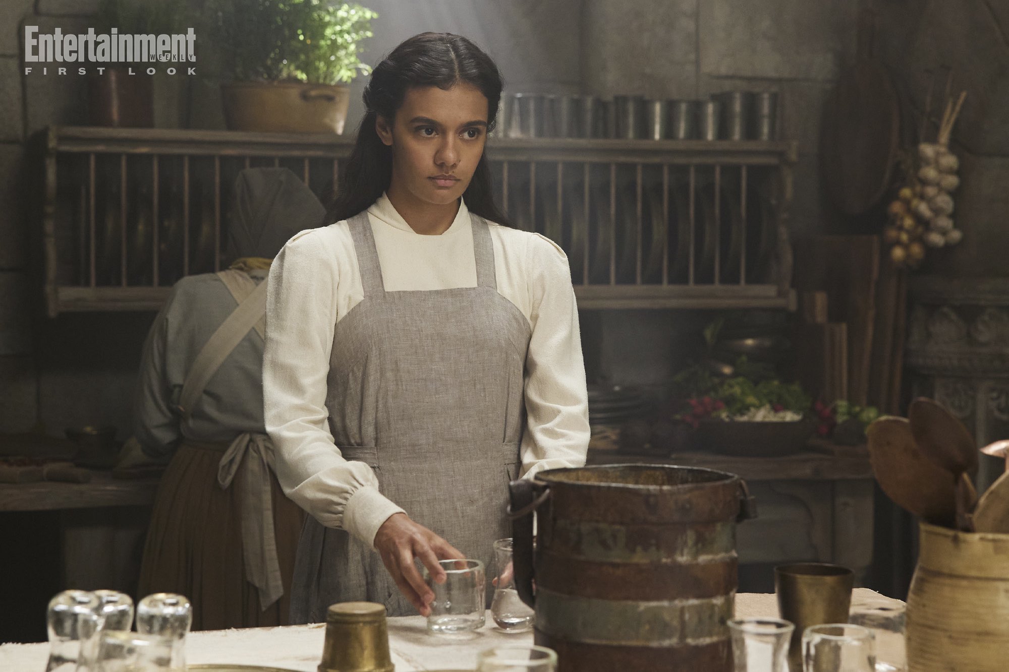 Egwene setting a table in the White Tower, dressed in novice white and a gray apron while Laras (?) cooks in the background