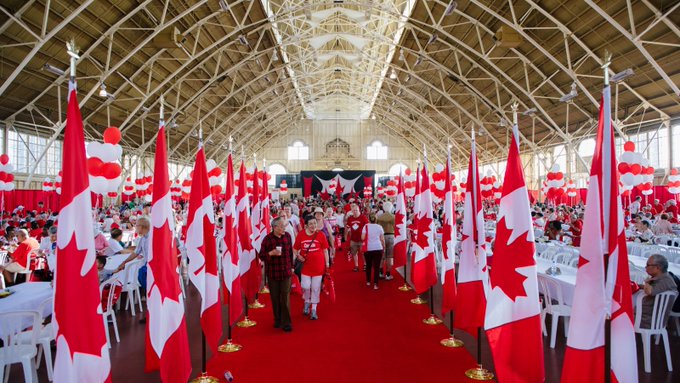 Canada Day flags line the Aberdeen Pavilion at Lansdowne Park. People mingle around a stage in the background. 