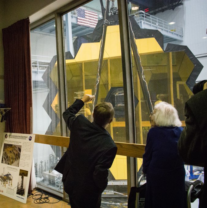 Dr. Jane Rigby and Dr. Nancy Grace Roman stand in front of the Webb telescope's golden hexagonal mirror, visible through a window that peers into the NASA Goddard cleanroom. Dr. Rigby has short blonde hair and wears a black suit. Dr. Roman has shoulder-length white hair and is wearing glasses and a dark blue dress.