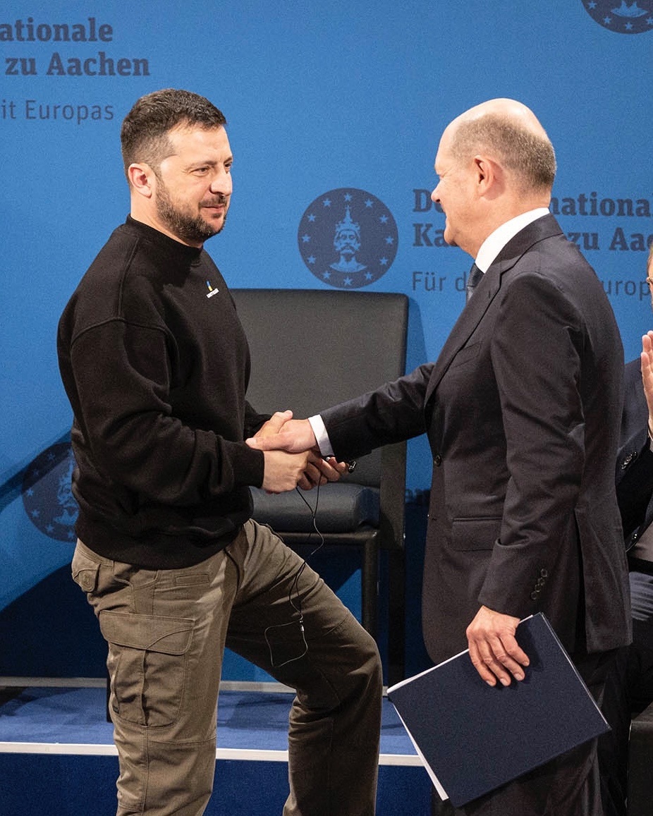 Das Foto zeigt den ukrainischen Präsidenten Wolodymyr Selensky und Bundeskanzler Olaf Scholz bei der Verleihung des Karlspreises in Aachen, nachdem Bundeskanzler Olaf Scholz die Laudatio gehalten hat. Beide stehen auf der Bühne und schütteln sich die Hand. Bundeskanzler Olaf Scholz hält eine blaue Umschlagmappe in der Hand. Im Hintergrund ist eine blaue Wand mit dem Emblem des Karlspreises zu sehen. 