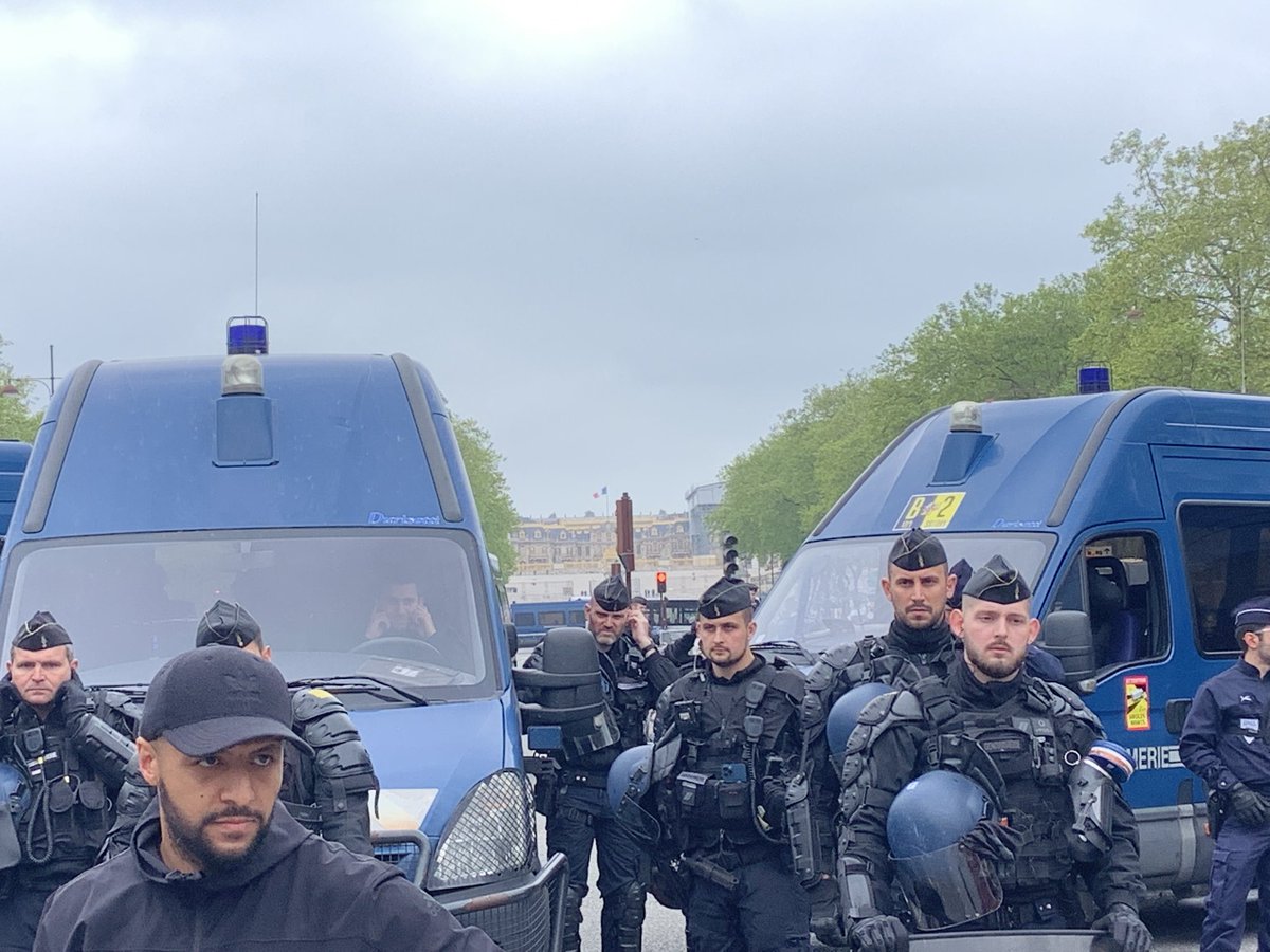 French workers marching w torches toward Versailles (like the peasants in 1789 revolution who deposed Louis 16th) where “king” Macron is holding an investment conference. Riot police hold them back. Palace in background.