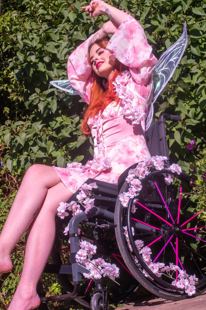ru, a white femme disabled person in their wheelchair. they are wearing a floaty pink and white dress with iridescent fairy wings. her wheelchair is decorated in pink cherry blossoms. their arms are above their head and she has a wistful facial expression.