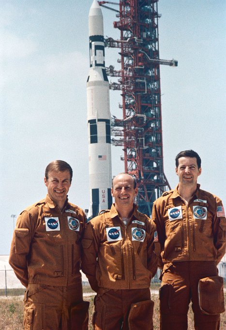 Paul Weitz, Pete Conrad, and Joseph Kerwin posing for a picture with the launch rocket for the Skylab 2 mission in the background.