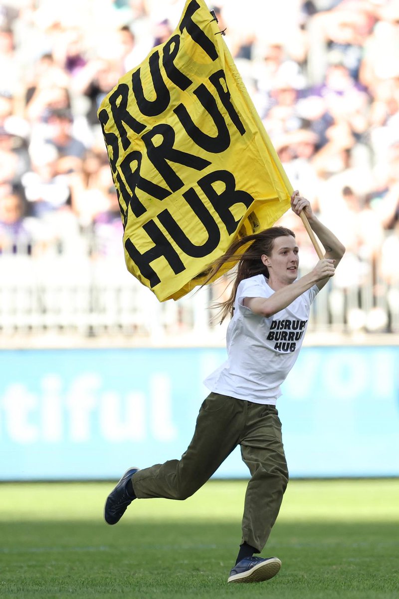 In the 2nd quarter of today’s #FreoVsCats game at @OptusStadium, musician & WAAPA student Emil Davey jumped over Woodside hoarding and onto the pitch with a DBH flag – disrupting play for several minutes – to demand no industry on the Burrup. #KalyakoorlWalyalup
